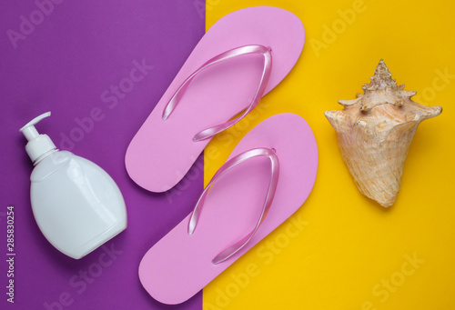 Beach accessories. Fashionable beach pink flip flops, sunblock bottle, seashell on purple yellow paper background. Flat lay. Top view