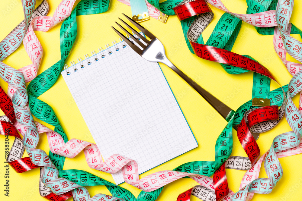 Fototapeta premium Top view of notebook with fork surrounded with colorful measuring tapes on yellow background. Copy space with diet planning
