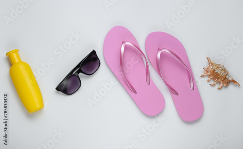 Summer still life. Beach accessories. Fashionable beach pink flip flops, sunblock bottle, sunglasses, seashell on white paper background. Flat lay. Top view
