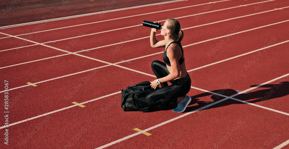 Obraz premium Young woman athlete in sportwear getting ready for workout while opening sports bag and drink water on the track of stadium at bright sunny day
