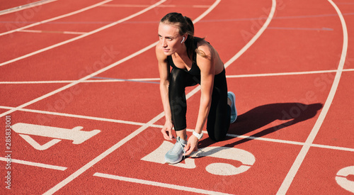 Young sprinter woman in sportswear tying shoelaces before a race on a red-coated stadium track at sunny bright day © splitov27