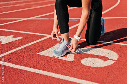 Young sprinter woman in sportswear tying shoelaces before a race on a red-coated stadium track at sunny bright day