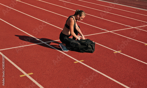 Young woman athlete in sportwear getting ready for workout and opening sports bag on the track of stadium at bright sunny day