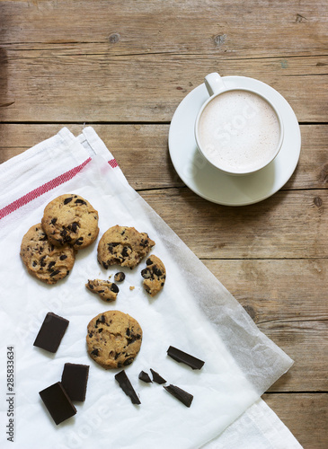 Chocolate cookies, pieces of chocolate and a cup of cocoa or coffee with milk on a wooden background. Rustic style.
