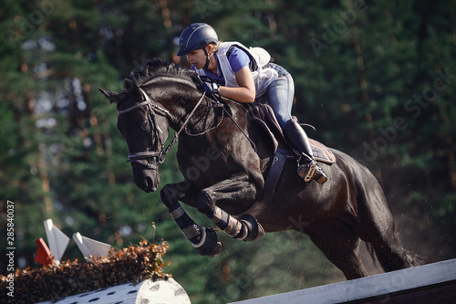 close portrait of attractive rider woman jumping over obstacle on black horse during eventing cross country competition in summer