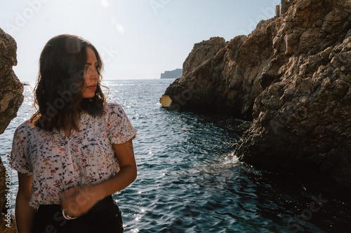 Adventurous young latin woman looking sideways on the sea rocks. Woman on cliff, with the sunset. Jovial near the sea on vacation in the Mediterranean with turquoise blue water.