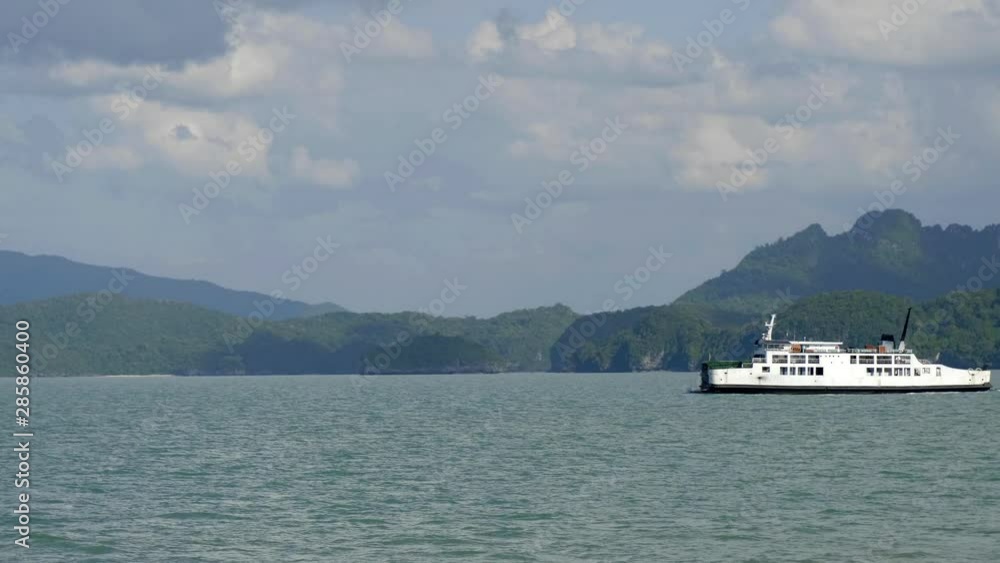 In a bay surrounded by lush green mountains, a large ferry sails into the frame. In a bay surrounded by lush green mountains, a large ferry sails into the frame.