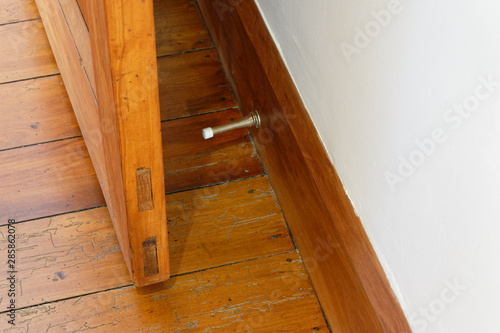 A wooden door is prevented from hitting a white wall by the metal spring door stopper that is attached to the wooden skirting board