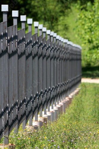 Dense row of wrought iron fence made of vertical metal poles with wrought iron cube on top mounted on concrete foundation surrounded with uncut grass and trees on warm sunny summer day