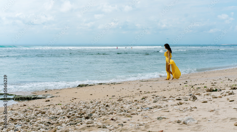 Obraz premium Woman in long yellow dress on rocky beach on Bali, Indonesia. Summer time. Dark hair brunette