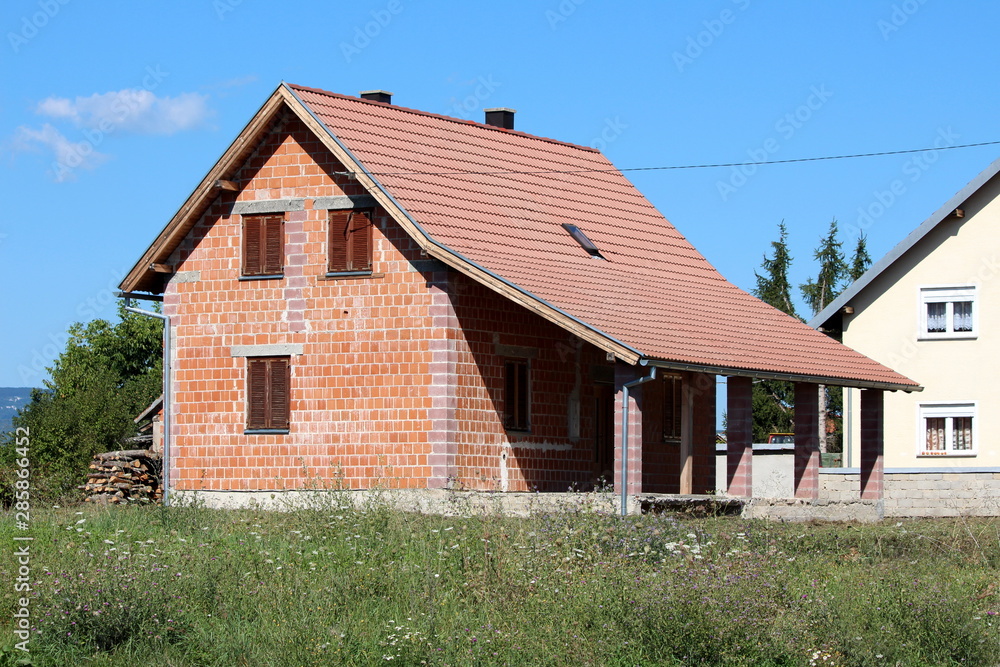 New unfinished small red brick family house with front porch surrounded with uncut grass and other houses on warm sunny summer day New unfinished small red brick family house with front porch surrounded with uncut grass and other houses on warm sunny summer day