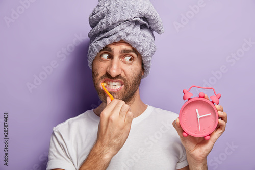 Handsome man brushes teeth, whitens with toothpaste, holds alarm clock in hand, awoke late in morning, has wrapped towel on head, wears casual white t shirt, isolated on purple wall. Morning routine