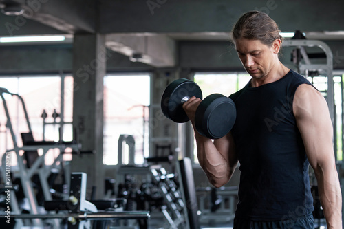 Sport man bodybuilder do workout exercise at fitness gym. Attractive muscular man in black sportswear standing do weightlifting dumbbells for strength and firm muscular. Healthy lifestyle concept.