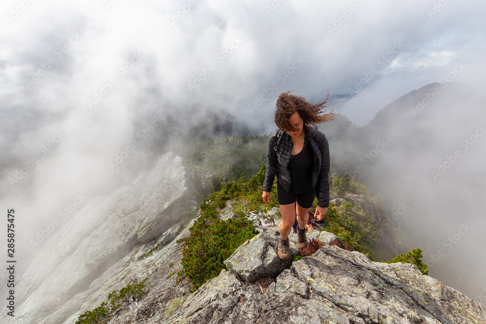 Naklejka premium Adventurous Girl is hiking up a steep trail up a beautiful rocky mountain during a cloudy summer morning. Taken on Crown Mountain, North Vancouver, BC, Canada.