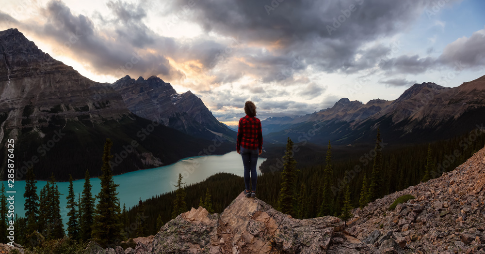 Adventurous girl standing on the edge of a cliff overlooking the beautiful Canadian Rockies and Peyto Lake during a vibrant summer sunset. Taken in Banff National Park, Alberta, Canada.