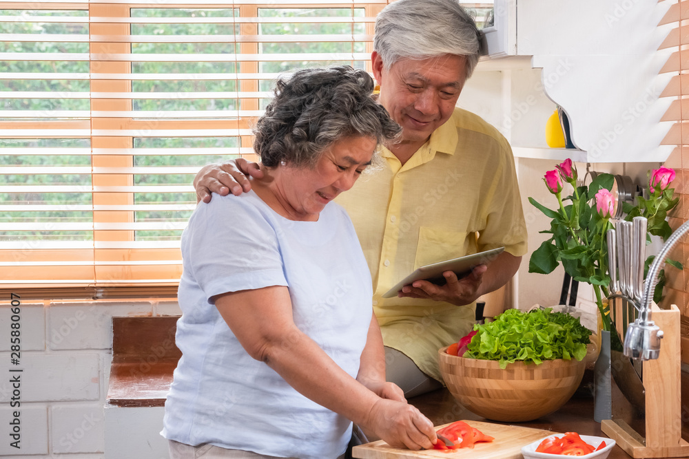 Asian senior wife slicing tomatoes on wooden cutting board and using tablet computer to searching menu recipe.senior with technology lfiestyle.aging at home Asian senior wife slicing tomatoes on wooden cutting board and using tablet computer to searching menu recipe.senior with technology lfiestyle.aging at home