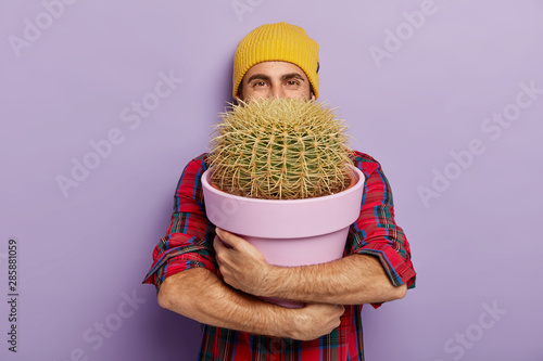 Photo of happy young male flower grower embraces big pot with prickly cactus, wears stylish hat and checkered shirt, glad to receive house plant as gift, isolated on purple wall. Gardening concept photo