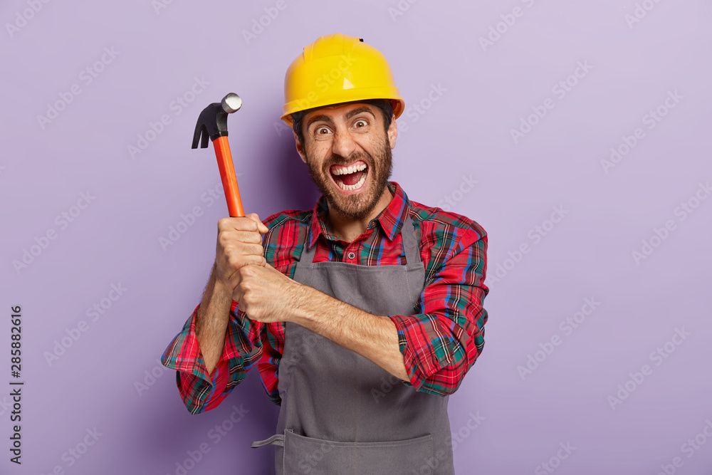 Desperate male laborer or repairman holds hammer with both hands, has outraged facial expression, ready for repairing or building, wears protective helmet, works at construction site, stands indoor. Desperate male laborer or repairman holds hammer with both hands, has outraged facial expression, ready for repairing or building, wears protective helmet, works at construction site, stands indoor.
