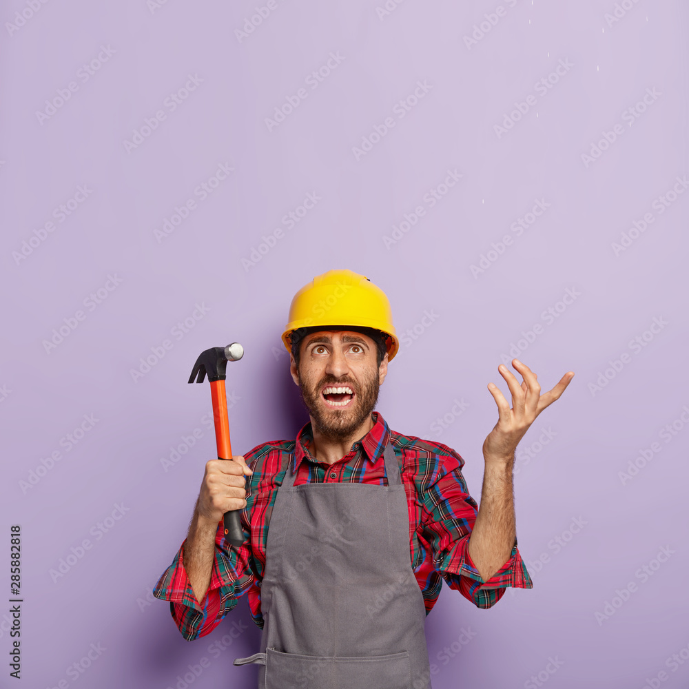 Unhappy handyman holds hammer, focused above with annoyance, repairs something with building tool in workshop, wears hardhat, shirt and apron. Foreman inspector at work, does repairing alone Unhappy handyman holds hammer, focused above with annoyance, repairs something with building tool in workshop, wears hardhat, shirt and apron. Foreman inspector at work, does repairing alone
