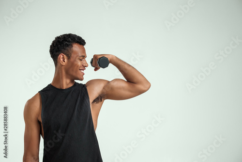 Making perfect body. Muscular young african man exercising with dumbbells and smiling while standing against grey background