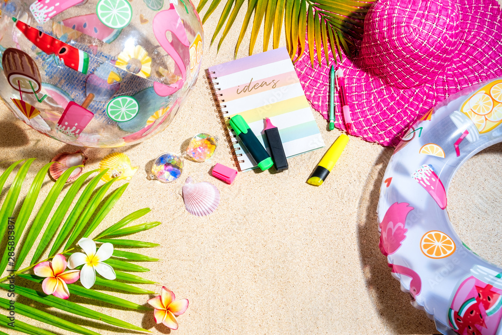 Fototapeta premium Colourful writing note pad with pink and green markers on sand, surrounded by green palm leafs, sea shells, pink hat, inflatable toys. Summer beach background, flat lay
