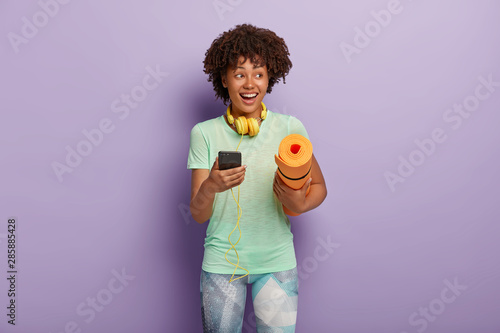 Horizontal shot of happy curly fitness woman listens music via headphones and smartphone during workout, carries rolled up karemat, dressed in t shirt and leggings. People, exercising concept