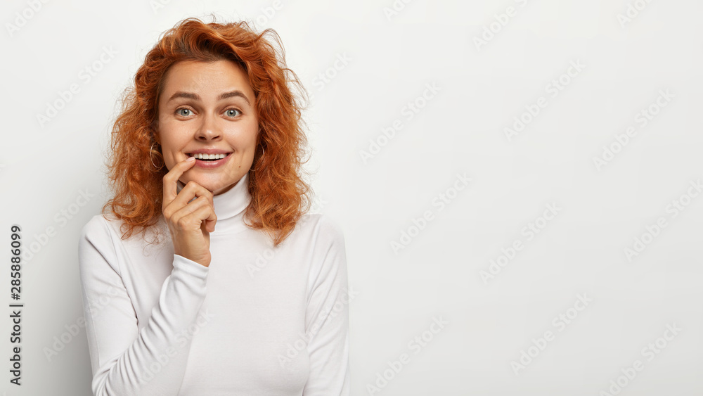 Portrait of happy gentle woman with red natural hair, holds index finger on lip, looks with great pleasure at camera, notices nice surprise, dressed casually, isolated on white background. Monochrome Portrait of happy gentle woman with red natural hair, holds index finger on lip, looks with great pleasure at camera, notices nice surprise, dressed casually, isolated on white background. Monochrome