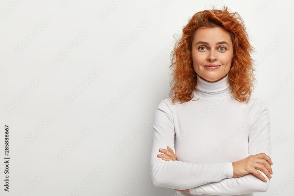 Portrait of good looking redhead woman looks with little smile, has calm face expression, keeps arms folded, wears white turtleneck, poses alone, listens pleasant comments, isolated on white wall Portrait of good looking redhead woman looks with little smile, has calm face expression, keeps arms folded, wears white turtleneck, poses alone, listens pleasant comments, isolated on white wall