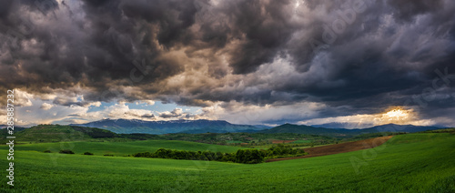 Panoramic view of uneven green agriculture fields in the foreground, distant mountain range in the background, and heavy stormy overcast dramatic clouds in the sky