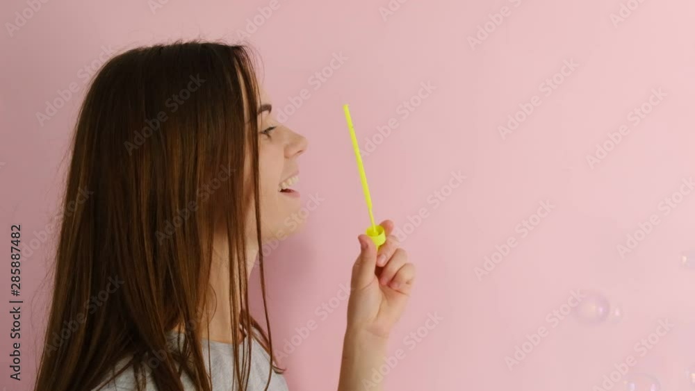 A beautiful cheerful young woman, wearing a t-shirt, standing isolated over pink background, smiling and blowing soap bubbles. People and emotion concept A beautiful cheerful young woman, wearing a t-shirt, standing isolated over pink background, smiling and blowing soap bubbles. People and emotion concept