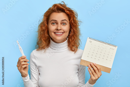 Glad foxy woman holds positive pregnancy test and ovulation calendar, controls women health, happy to find out about future maternity, wears white sweater, isolated on blue wall. Pregnancy planning