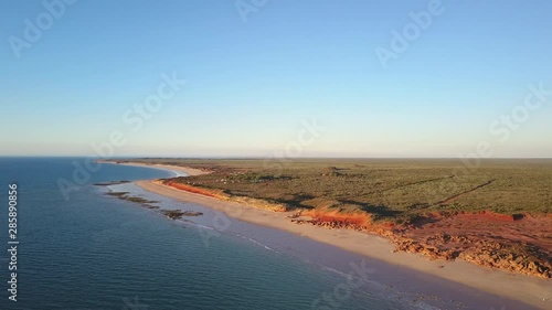 Scenic panoramic aerial drone view of camping area at remote beach in Western Australia, flying along ocean, red rocky cliffs, with blue sky, sunset and horizon on a summer sunny day.