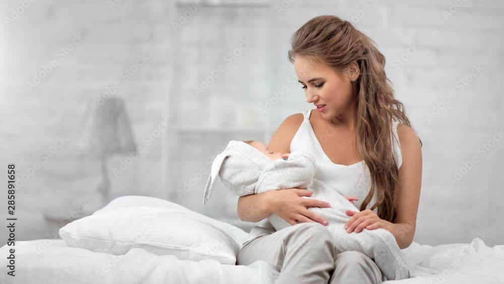 Caring enthusiastic pretty mother lulling sleeping baby sitting on bed at white room medium shot Caring enthusiastic pretty mother lulling sleeping baby sitting on bed at white room medium shot