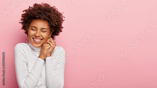 Horizontal shot of lovely dark skinned teenage girl laughs and expresses sincere emotions, keeps hands together near face, being in high spirit, feels overjoyed isolated on rosy wall free space aside