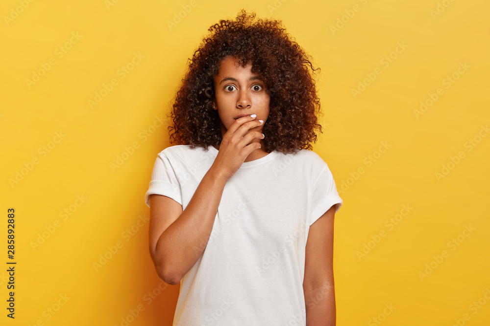Scared emotional dark skinned girl with luminous curly hair, covers face and looks with bugged eyes at camera, looks with terrified expression, frightened for own life, holds breath from excitement Scared emotional dark skinned girl with luminous curly hair, covers face and looks with bugged eyes at camera, looks with terrified expression, frightened for own life, holds breath from excitement