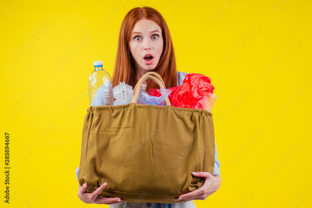 redhaired ginger european woman with cotton eco bag full of plastic trash in yellow studio background.recycling ecology solution concept redhaired ginger european woman with cotton eco bag full of plastic trash in yellow studio background.recycling ecology solution concept