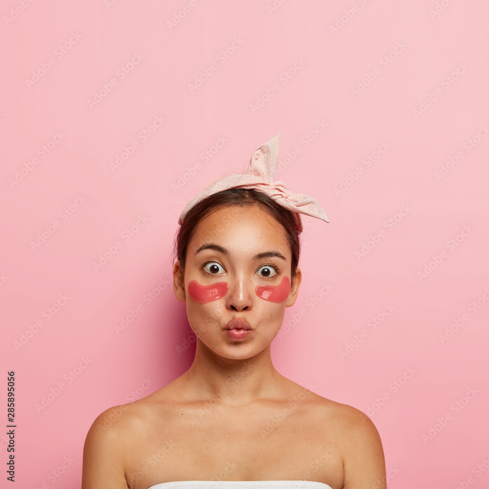 Adorable Asian woman applies self sticking patches under eyes to reduce puffiness and dark circles, wears headband on head, keeps lips folded, stands bare shoulders, isolated on pink background - obrazy, fototapety, plakaty 