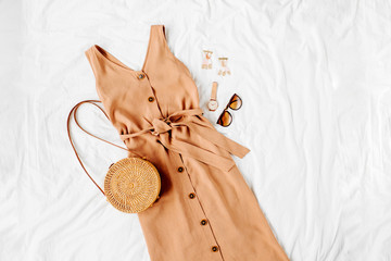 Beige dress with bamboo bag and accessories on white bed. Women's stylish autumn or summer outfit. Trendy clothes. Flat lay, top view.