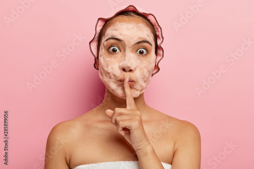 Image of surprised korean lady with soap bubbles on face, makes silence gesture, tells beauty secret, cleans and exfoliates skin, has cosmetic procedures during free time, takes care of herself photo