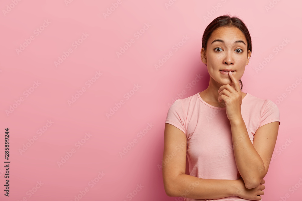 Photo of confused Asian girl bites lips and looks nervousy at camera, holds hands partly crossed, wears piercing in ear, dressed in casual t shirt, stands againt pink wall with blank space aside Photo of confused Asian girl bites lips and looks nervousy at camera, holds hands partly crossed, wears piercing in ear, dressed in casual t shirt, stands againt pink wall with blank space aside