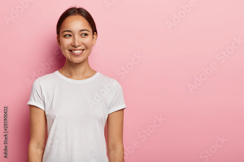 Obraz na plátně Studio shot of happy young Asian woman has tender smile, looks aside with charming expression, wears casual white t shirt, has natural beauty, isolated on pink wall