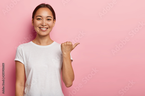 Horizontal shot of happy young Asian woman points away on copy space, demonstrates something good, wears white t shirt, helps pick best choice, recommends product, models over pink background
