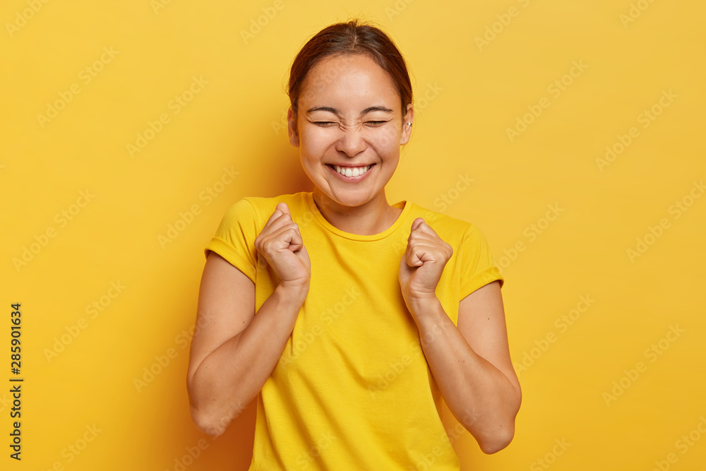 Yes, finally success! Joyful korean girl clenches fists with triumph, closes eyes from happiness and joy, has toothy smile, wears casual outfit, isolated on yellow background, triumphs victory Yes, finally success! Joyful korean girl clenches fists with triumph, closes eyes from happiness and joy, has toothy smile, wears casual outfit, isolated on yellow background, triumphs victory
