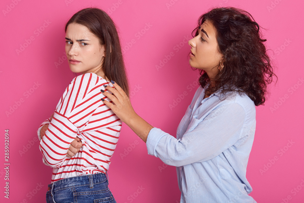 Beautiful girls with dark hair, lady wears striped shirt keep arms folded, do not talk to her friend, woman dressed blue shirt touches female's shoulders, wants to talk, pose isolated over pink wall. Beautiful girls with dark hair, lady wears striped shirt keep arms folded, do not talk to her friend, woman dressed blue shirt touches female's shoulders, wants to talk, pose isolated over pink wall.