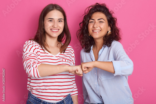 Smiling caucasian women fist bumping in studio, two friends greeting or supporting team motivation, females wear casual shirtts, have darl hair, looks at camera with toothy smile, expresse happyness. photo