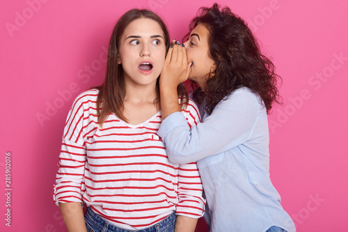 Two young attractive european girls wearing casual clothes standing and posing isolated over pink wall background, studio portrait. People lifestyle concept. Woman wispering secret to friend's ear. photo