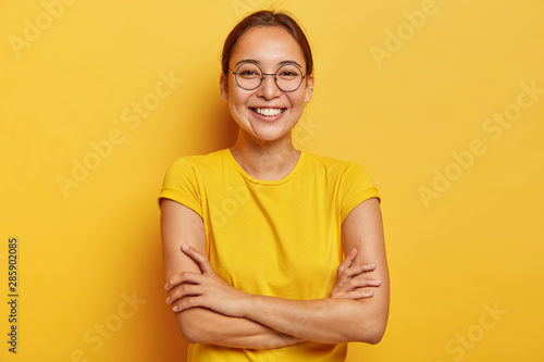 Isolated shot of pleased cheerful woman with eastern appearance, smiles broadly, being in good mood, entertained by funny friends, dressed casually, wears big transparent glasses, isolated on yellow