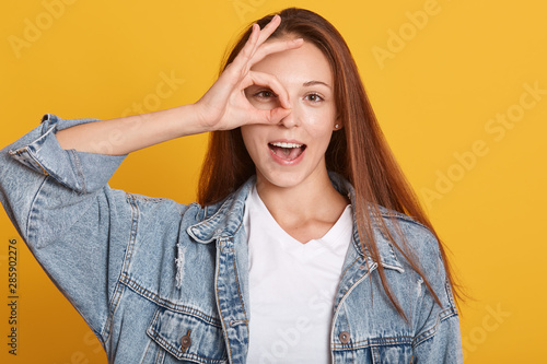 Studio shot of charming young woman, female making ok sign and covers her eye, posing with open mouth, expresses happyness, model posing over yellow background, being photographing in studio. photo