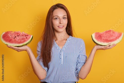 Photo of woman holding two pieces of watermelon in hands, looking directly at camera with charming smile, having long brown hair, wearing striped shirt, spreding hands, People and summertime concept. photo