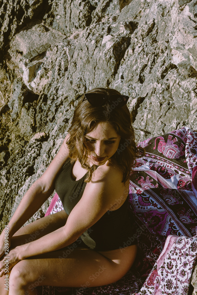 Naklejka premium Young hipster woman lying on the rocks in a swimsuit over beach towel. Young woman lying near the sea with vintage sunglasses.